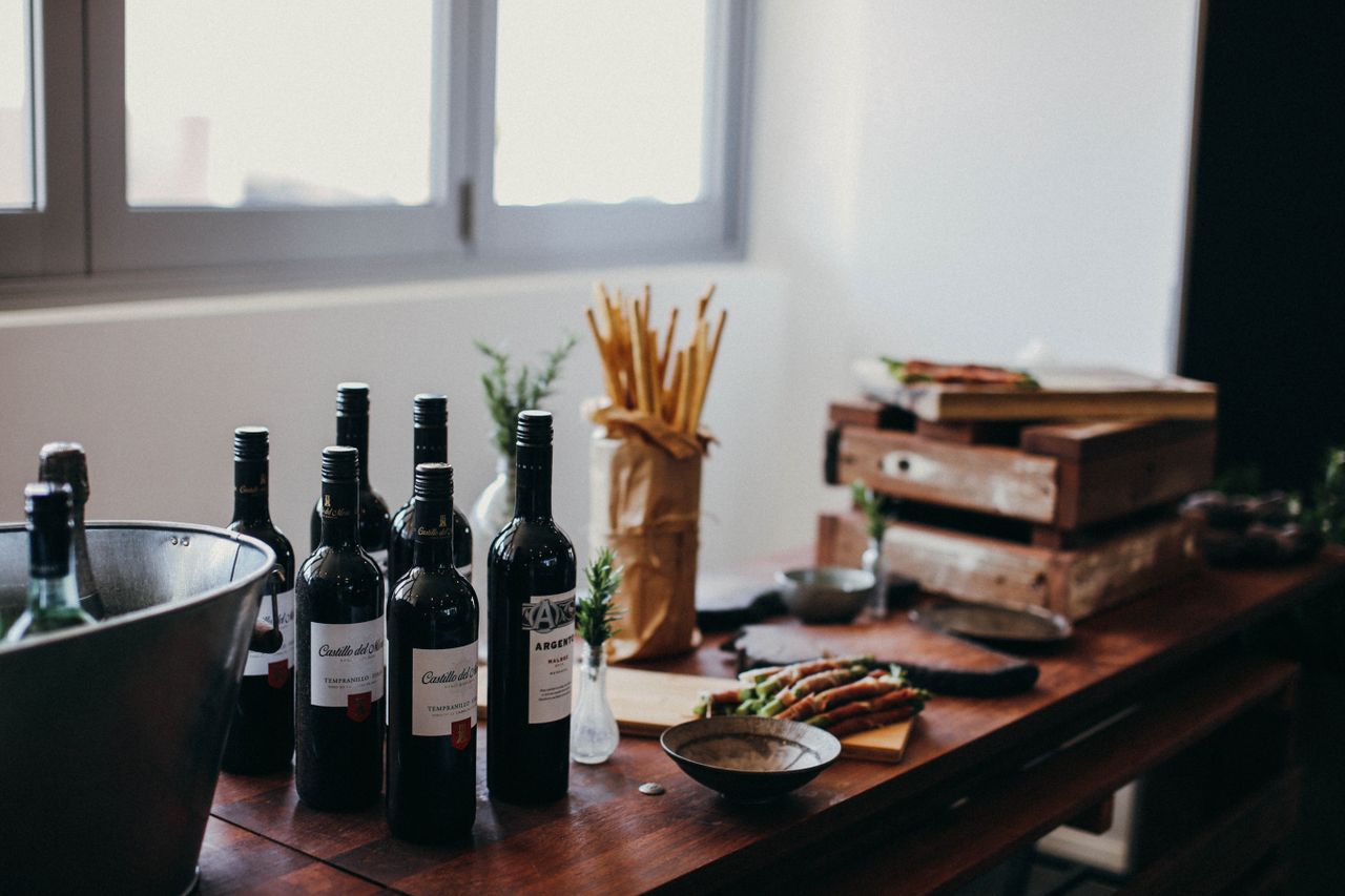 about-img Various bottles of alcoholic drinks placed on table with grissini sticks and wooden boxes near green branches in vase in room