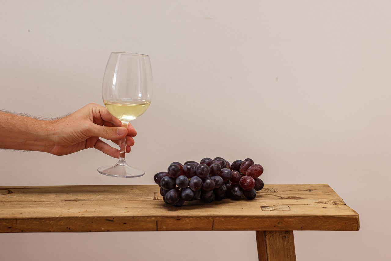 social-feed-img-05 A hand holds a white wine glass beside a bunch of grapes on a wooden bench, symbolizing elegance.